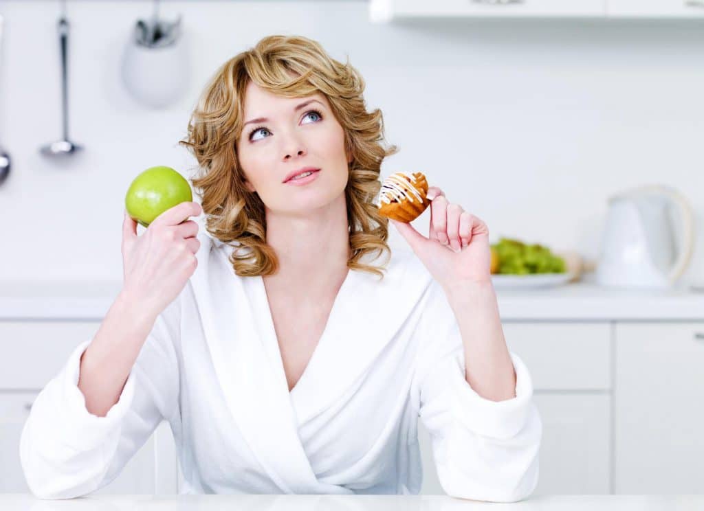 A woman at a breakfast table in a terrycloth robe tries to decide between eating an apple and eating a muffin for her morning meal. Apples are an excellent source of prebiotic fiber.