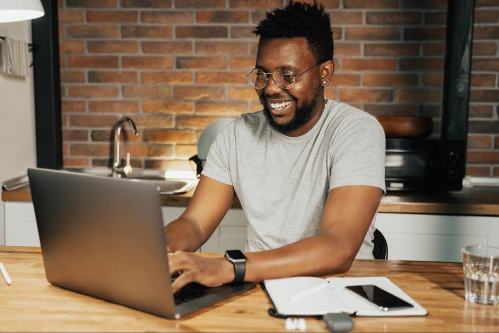 Black man smiling typing on laptop with notebook and phone beside him.