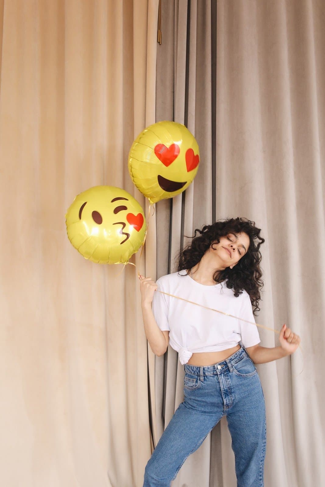 woman-in-white-shirt-and-blue-denim-pants-holding-yellow-emoji-balloons